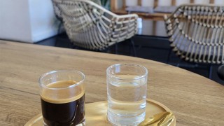 A small glass of espresso a small glass of water and a gold spoon on a wooden tray